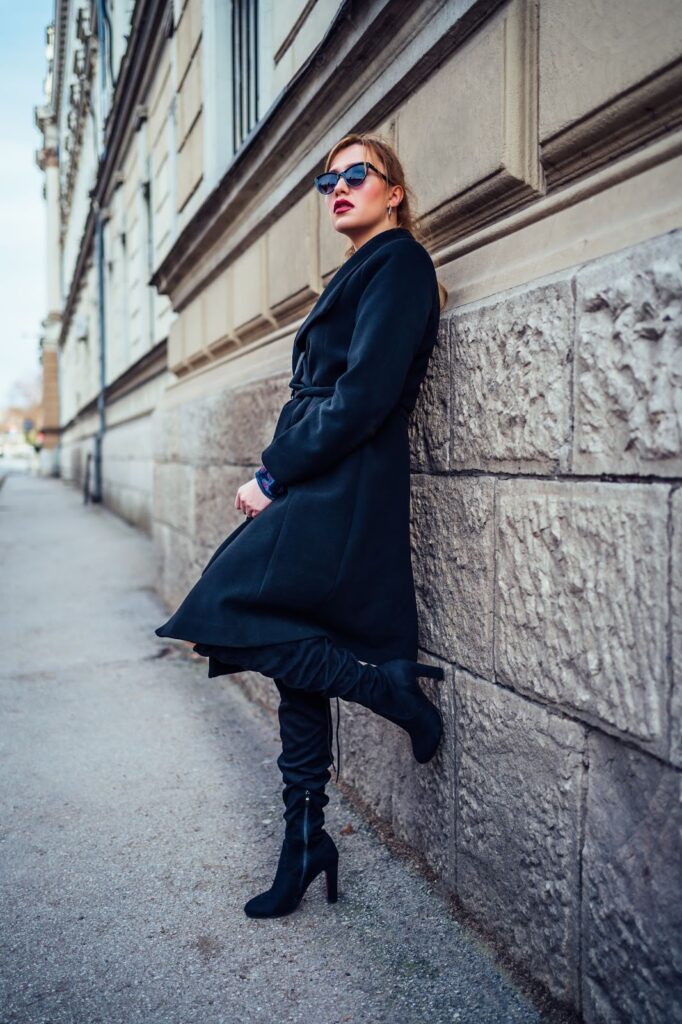 Жінка стоїть під стіною будинку у чорному пальті, чорних окулярах та високих чорних чоботах на каблуку.