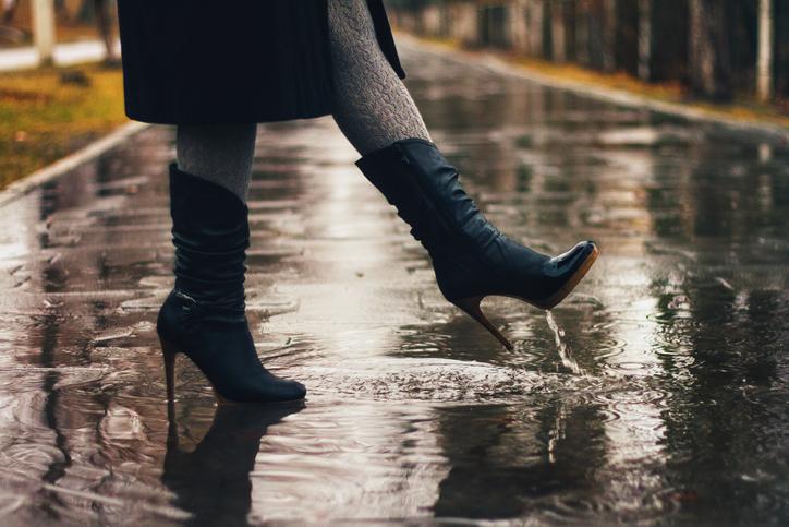 Жінка йде по мокрому асфальті у високих чоботах на шпильці.
