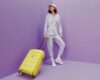 Дівчина із жовтою валізою стоїть на фіолетовому фоні.