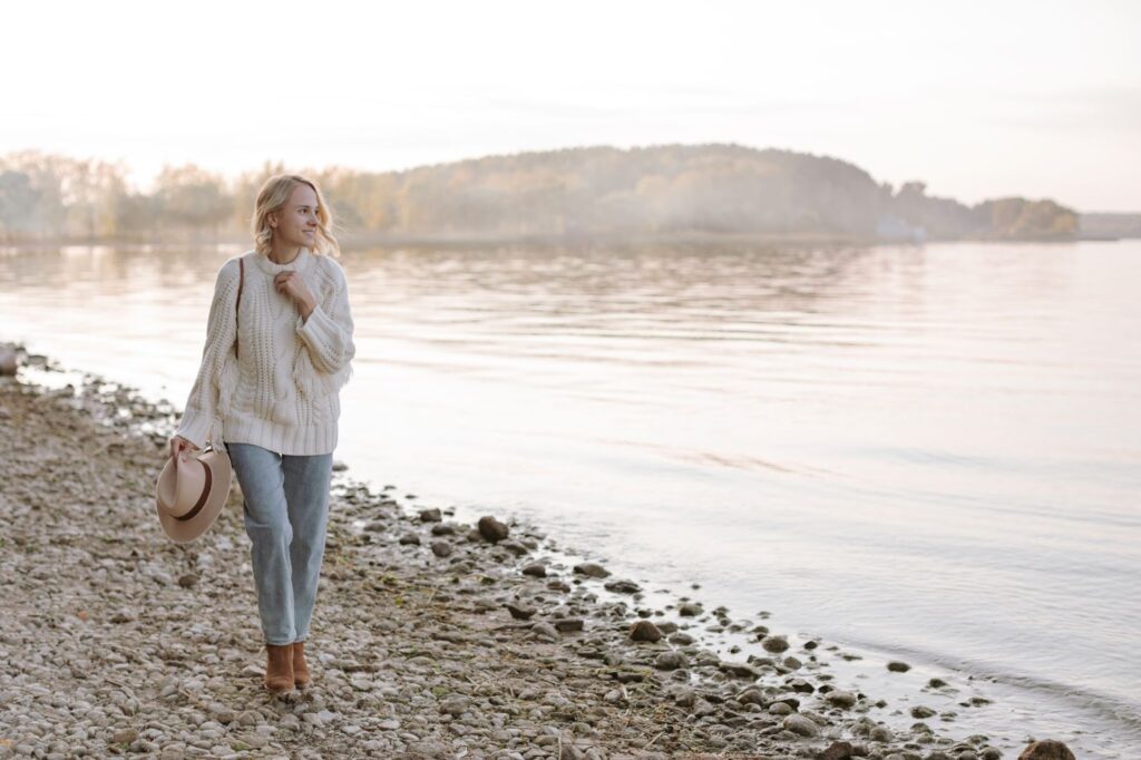 Дівчина йде на березі озера у джинсах, в’язаному светрі, коричневих ковбойських чоботах і тримає в руках капелюх бежевого кольору.