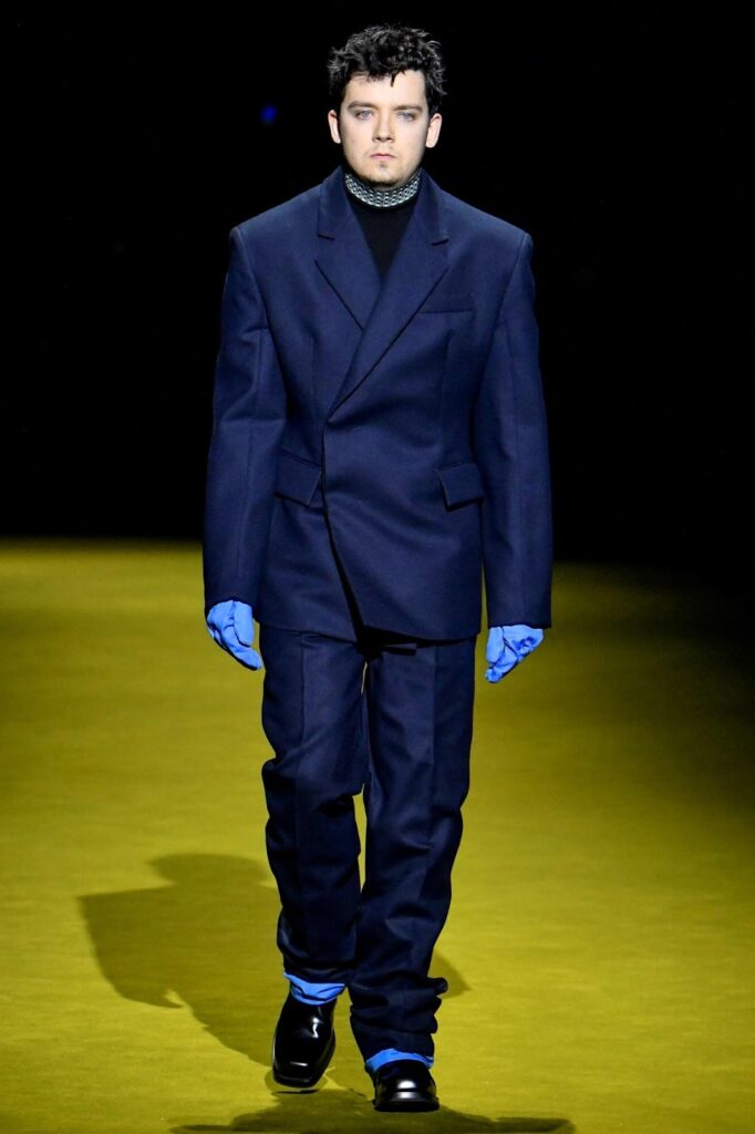 Модель йде по подіуму у синьому костюмі, блакитних рукавичках і чорних туфлях на товстій підошві.