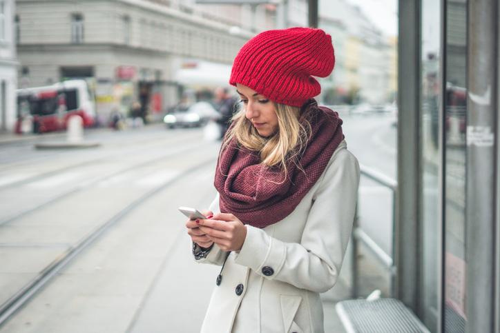 Дівчина стоїть біля трамвайної колії тримаючи в руках смартфон і одягнена в світле пальто в поєднанні з бордовим шарфом і червоною шапкою.