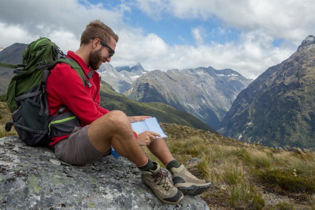 Чоловік сидить на камені в горах, одягнений у червоний спортивний джемпер, коричневі спортивні шорти, шкарпетки і трекінгові черевики.