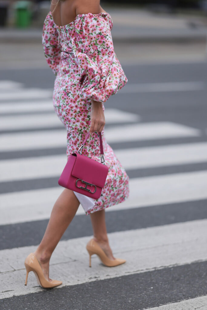 Жінка в квітковій сукні з відкритими плечима, яскраво-рожевою сумочкою та бежевими лакованими туфлями на шпильці йде по вулиці