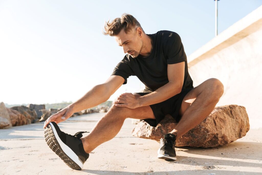 Чоловік, який робить розтяжку перед ранковою пробіжкою на пляжі в чорній футболці, шортах і чоловічому спортивному взутті