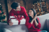 ідея подарунка для неї – чоловік дарує подарунок жінці, обидва в червоних светрах, святкова атмосфера навколо